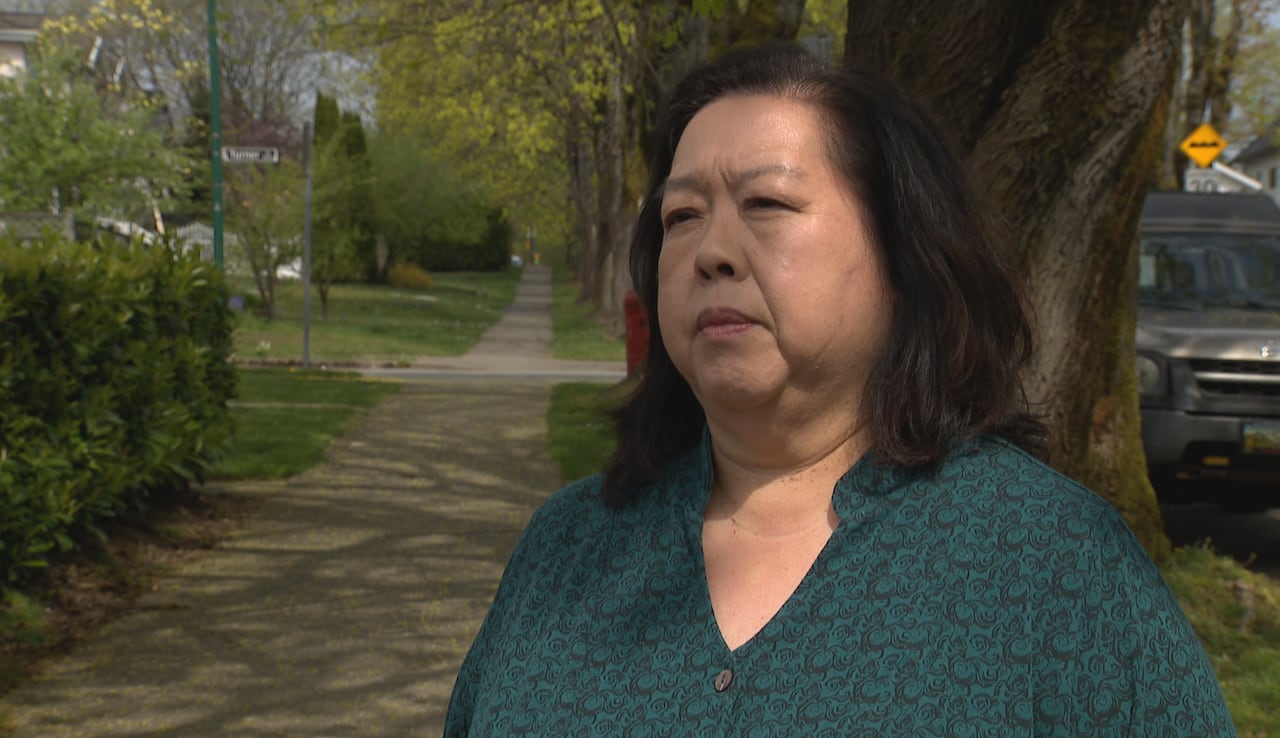 A woman with dark, shoulder-length hair speaks on a pavement next to trees.