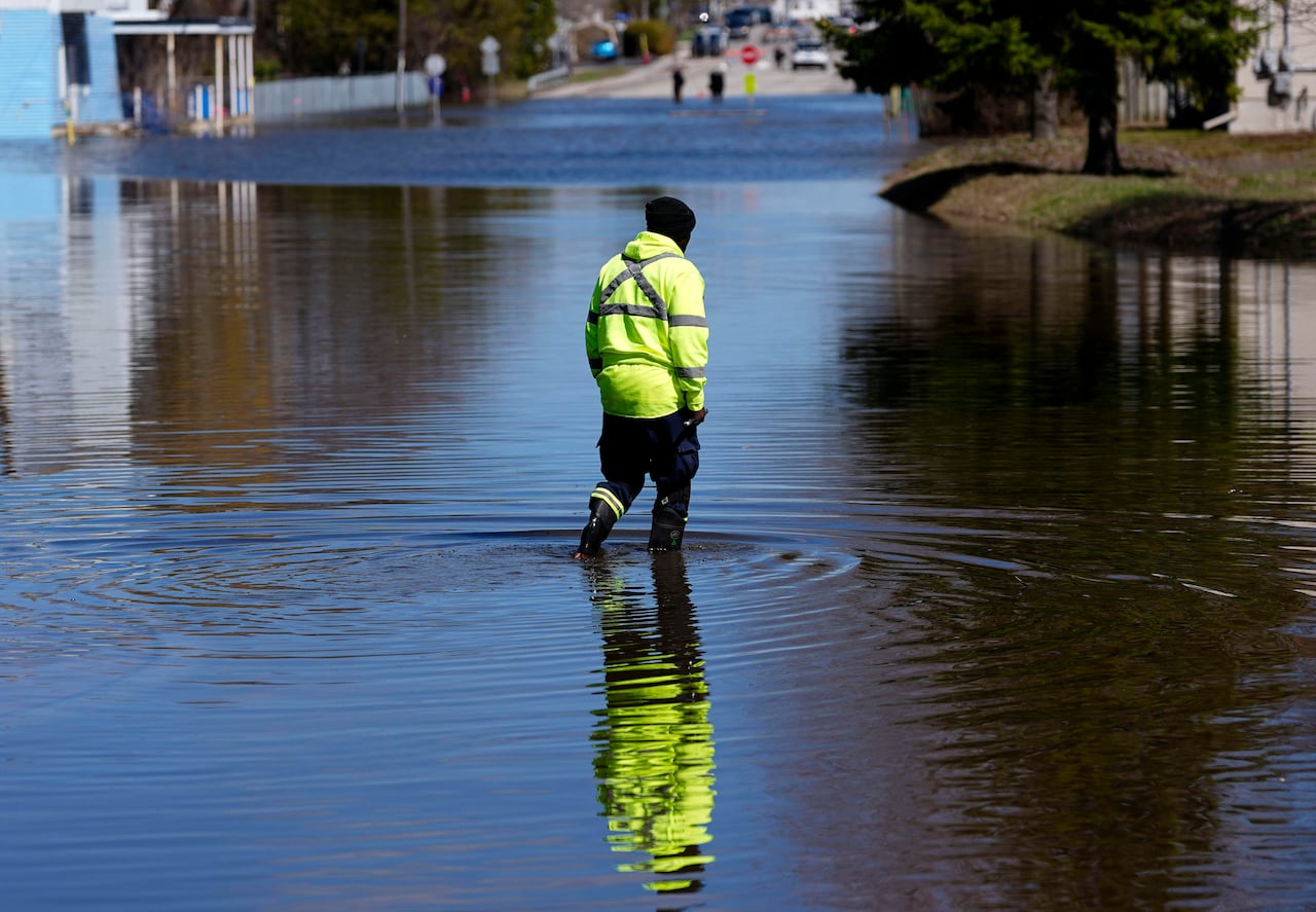A worker in a bright yellow jacket walks on a flooded city street on a sunny day.