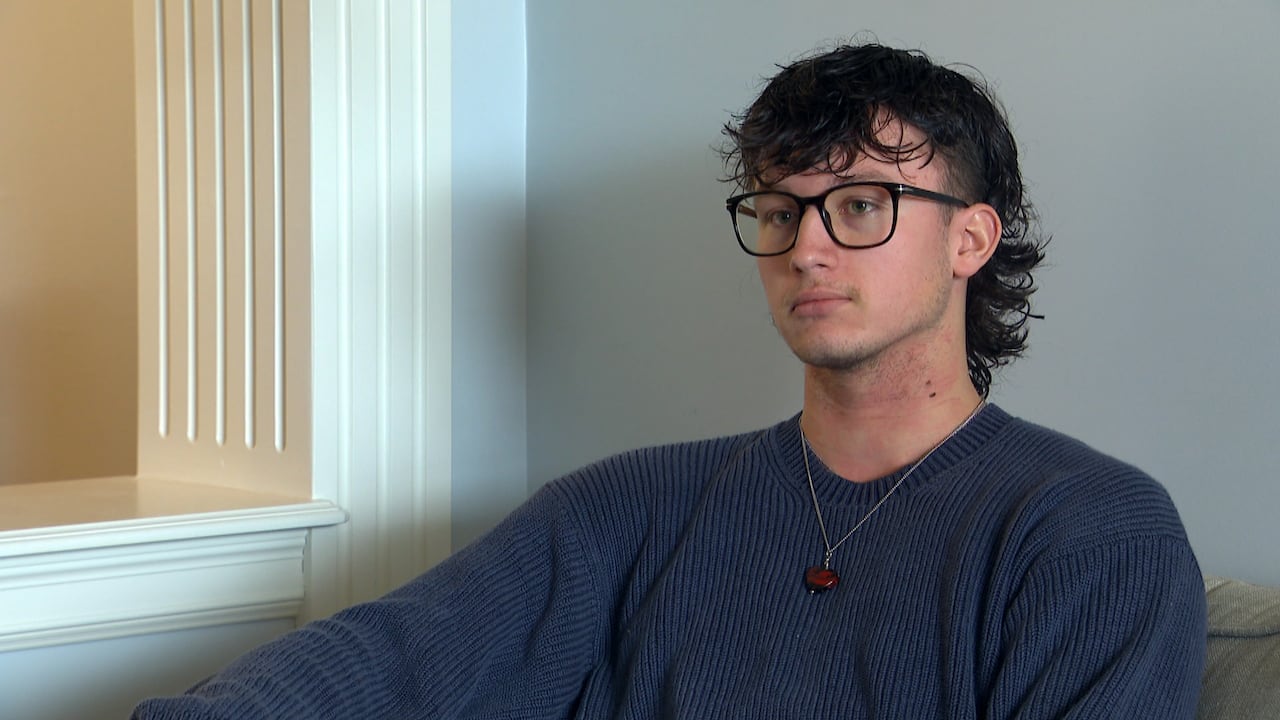A man with long brown hair sits on a couch. He's wearing black glasses and a navy blue sweater.