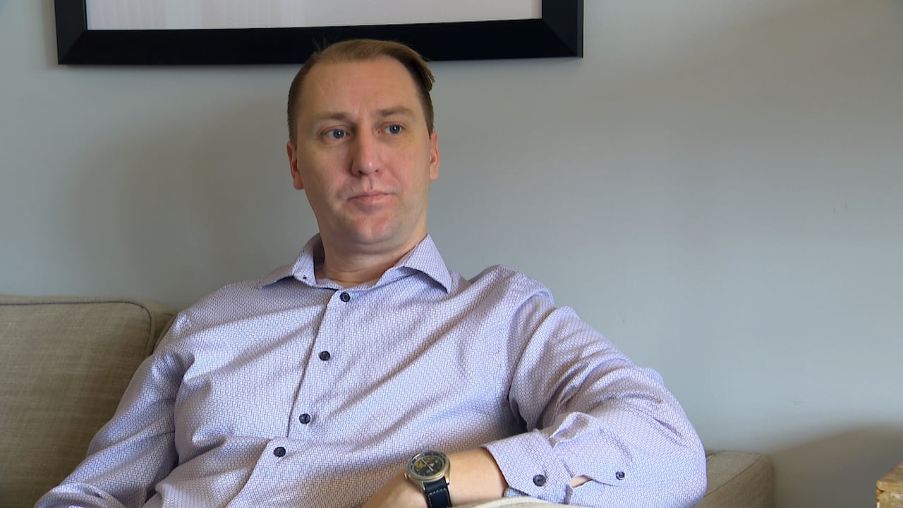 A man with short blonde hair sits on the couch. He is wearing a light purple button-up shirt and a black watch on his wrist.