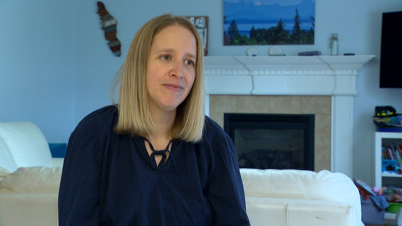A woman with blonde hair sits in front of her living room. She is wearing a navy blue blouse and there is an unlit fireplace behind her.