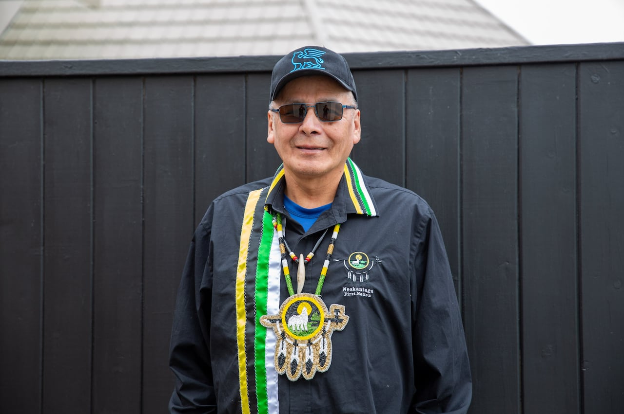 A person wearing a ribbon shirt and beaded medallion is seen standing in front of a black fence outside.