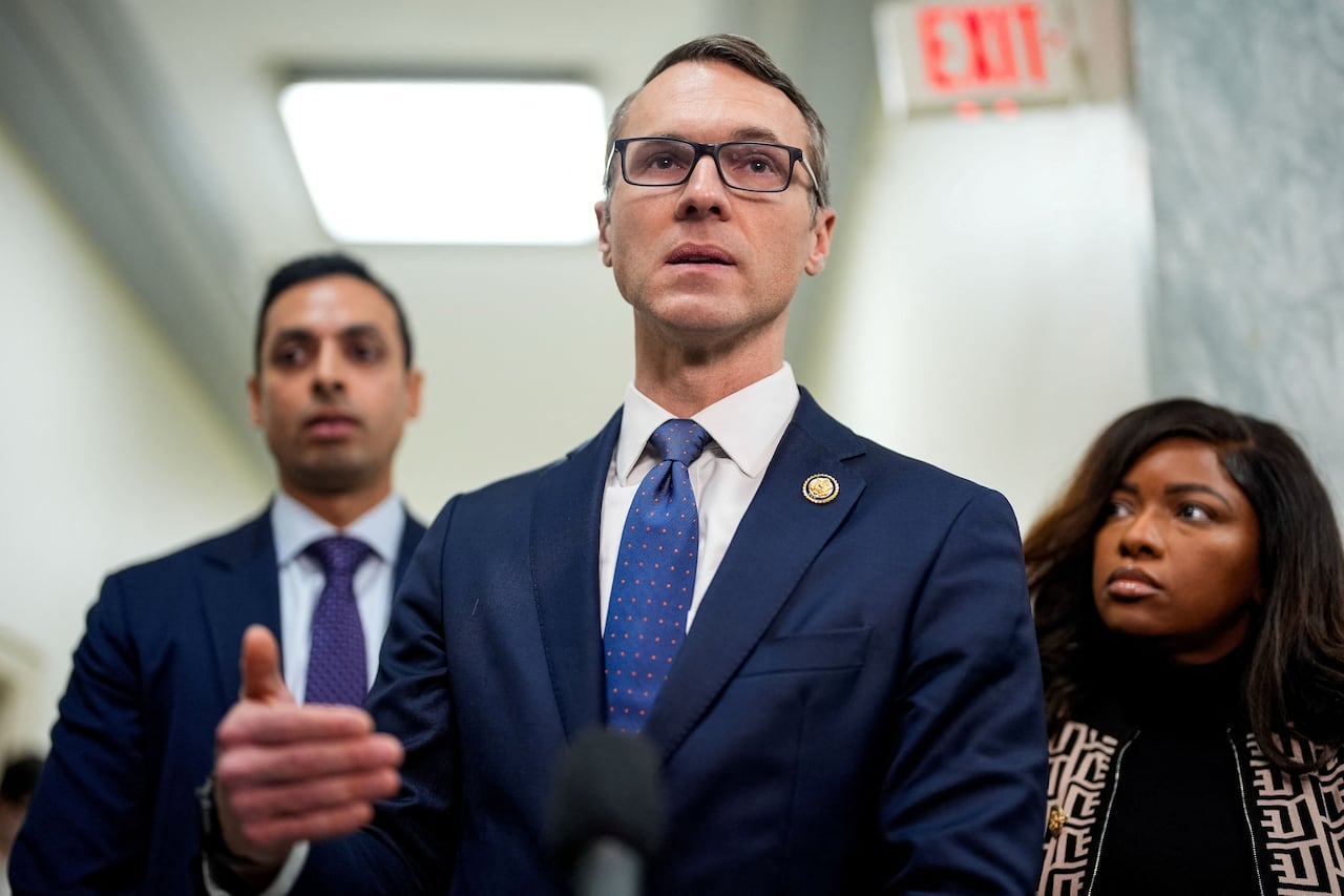 A bespectacled, cleanshaven man in suit and tie stands and speaks as a man and a woman stand near him.