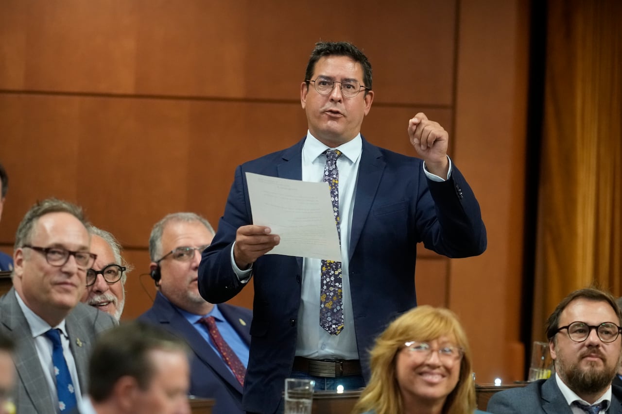 A man in a suit reads off a piece of paper and gestures with his hand. Others listen.