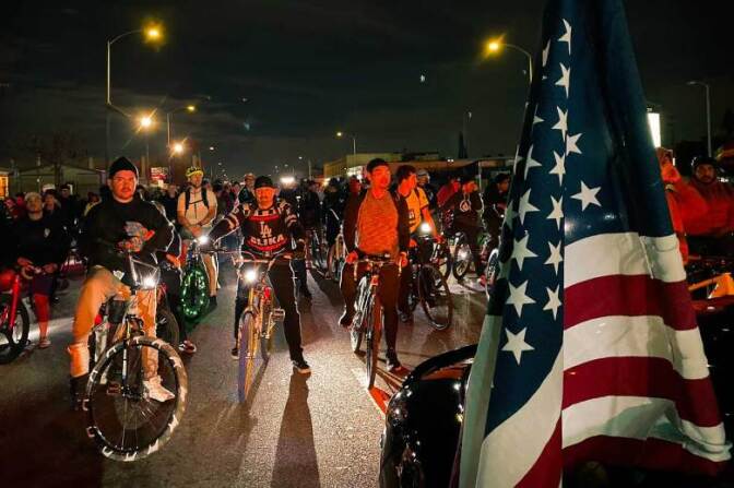 Thousands of bike riders stand around a street at night. An American flag is set up in the foreground by a car.