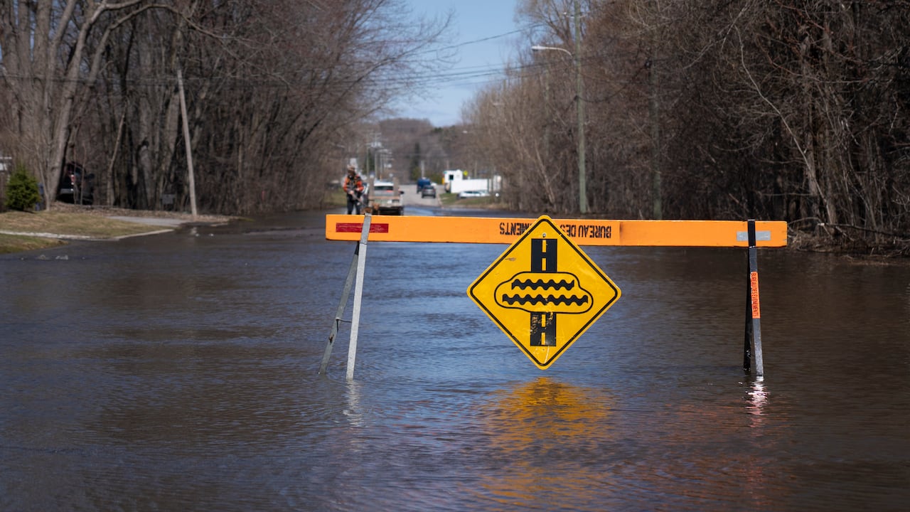 Low floodwaters cover a residential street on a spring day. There is a barricade in front of the flooded section.