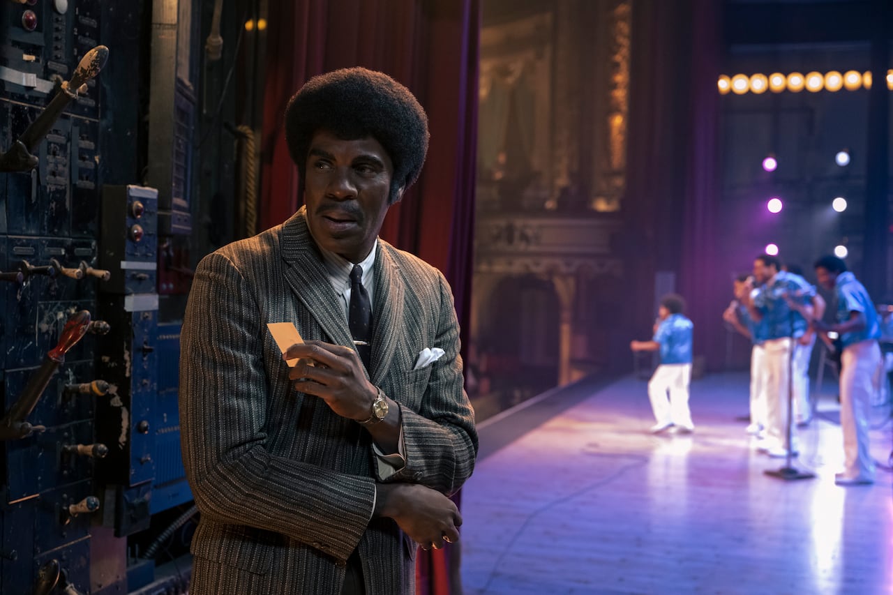 Aman in a suit stands in the wings of a theatre. Behind, a group of young performers can be seen onstage.