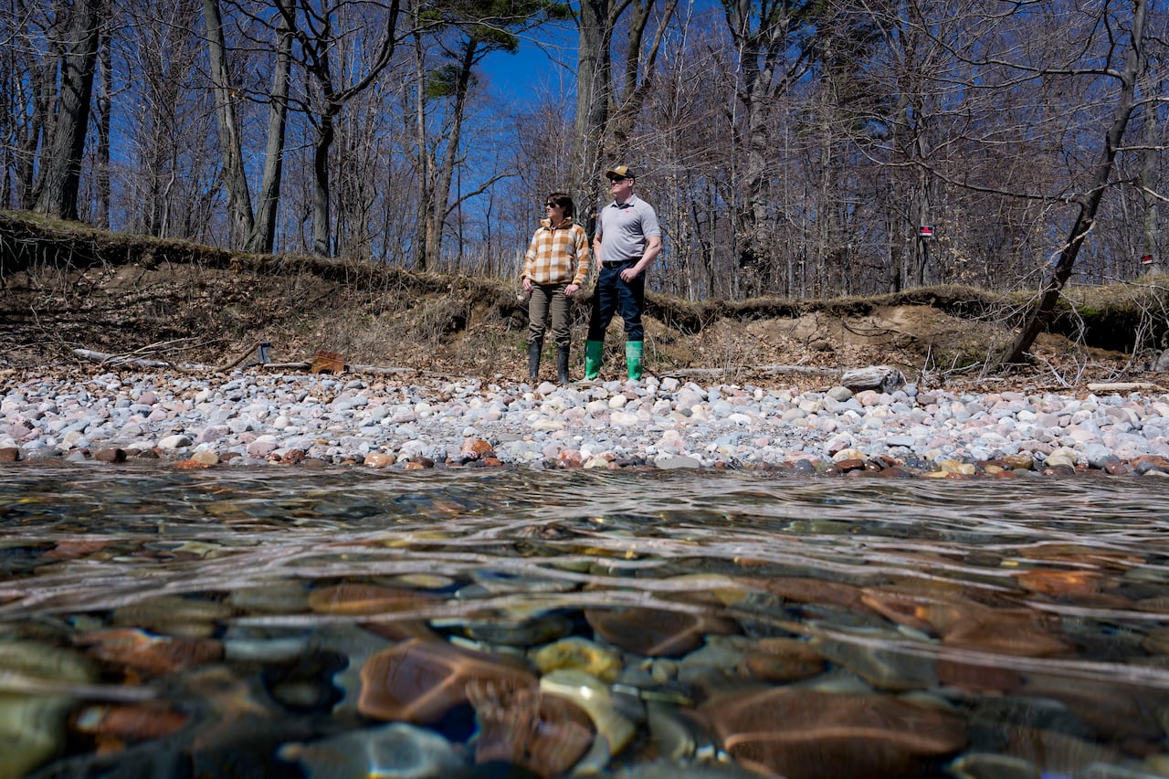 A white woman and white man both wearing sunglasses stand in front of a swath of small white rocks at the edge of a river. There are trees without leaves behind them.