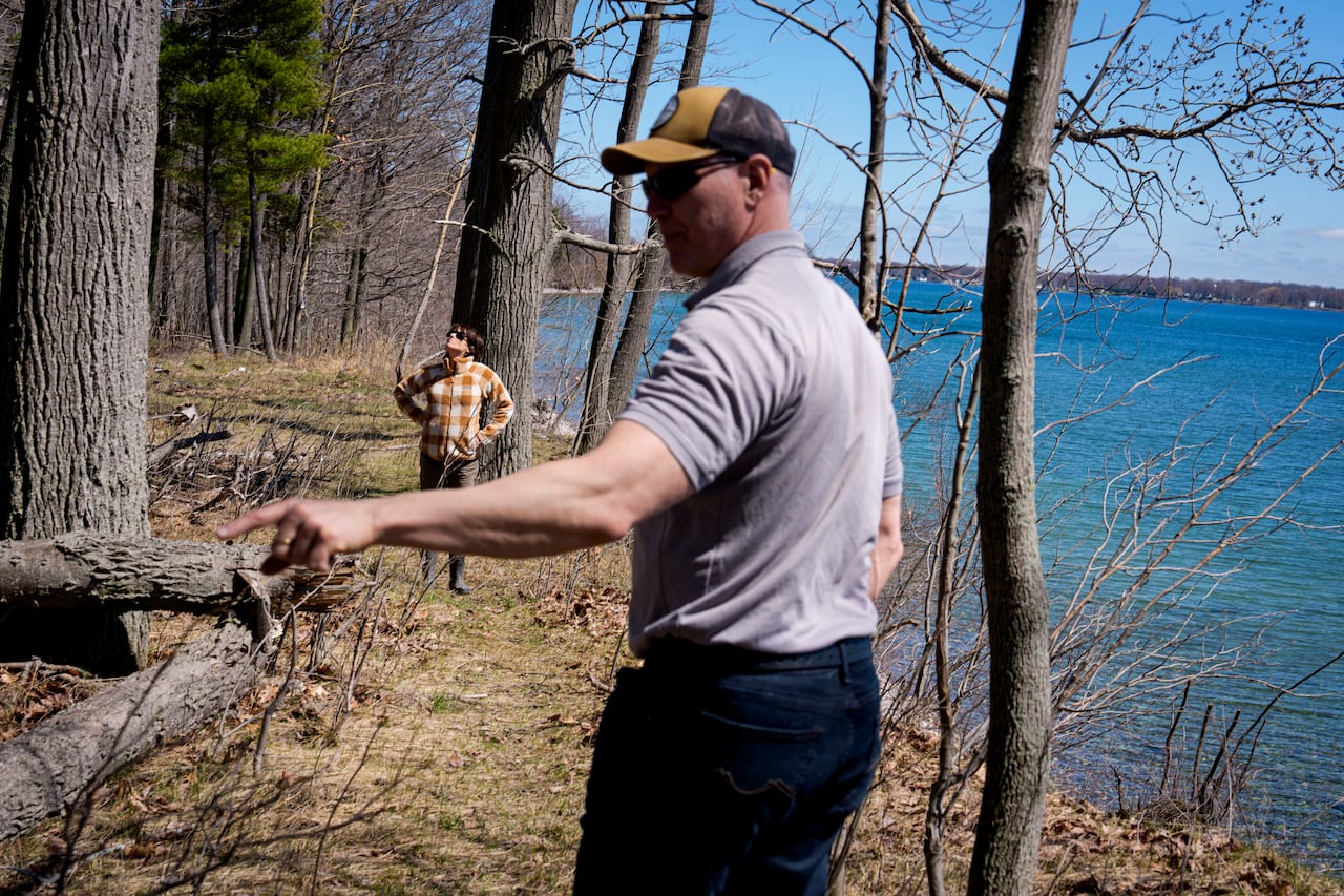 A white man wearing a gray shirt, yellow hat and sunglasses points to the left with his back to the camera. A white woman wearing sunglasses and a checkered jacket is in the background looking up.
