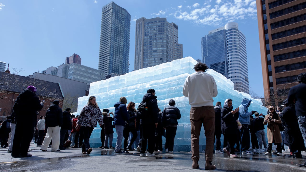 People around a large stack of ice blocks in a parking lot, with skyscrapers in the distance.