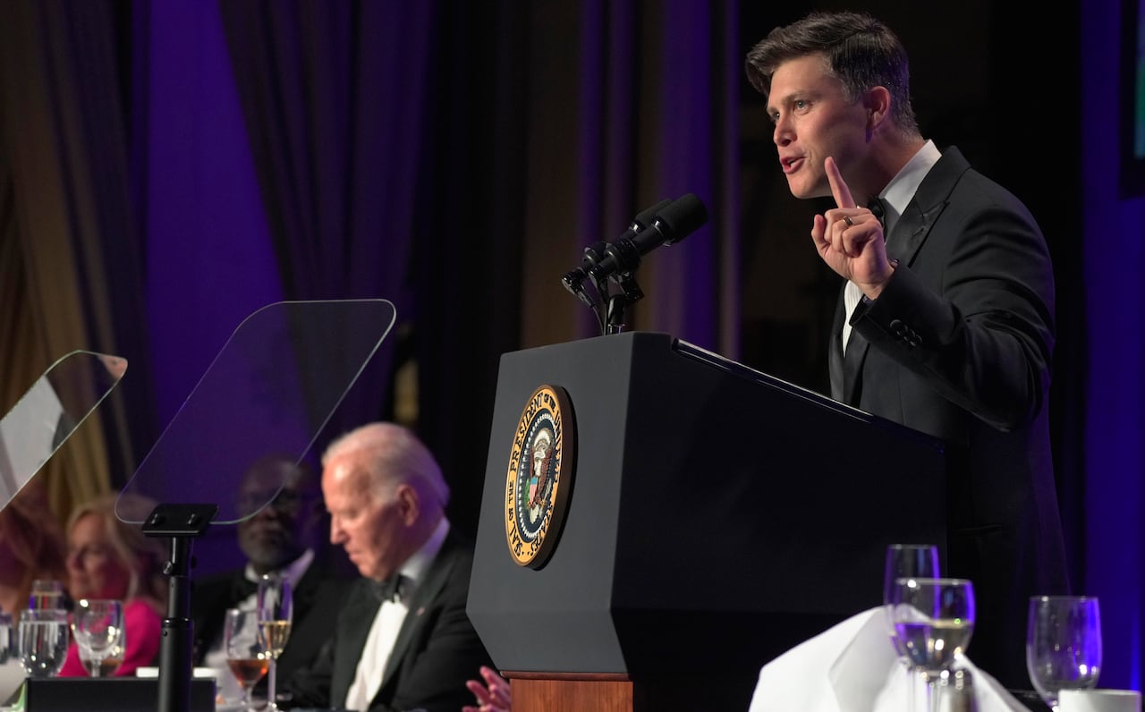 Colin Jost stands at a podium with the U.S. Presidential Seal, with Joe Biden seen in the background.