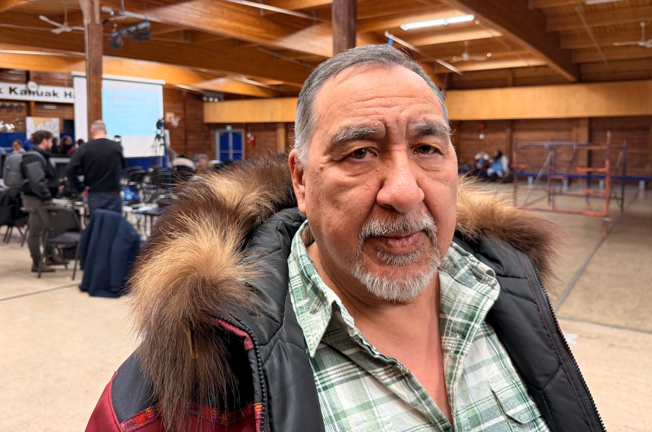 A grey-haired man wearing a winter parka and a plaid shirt is shown in a community hall.