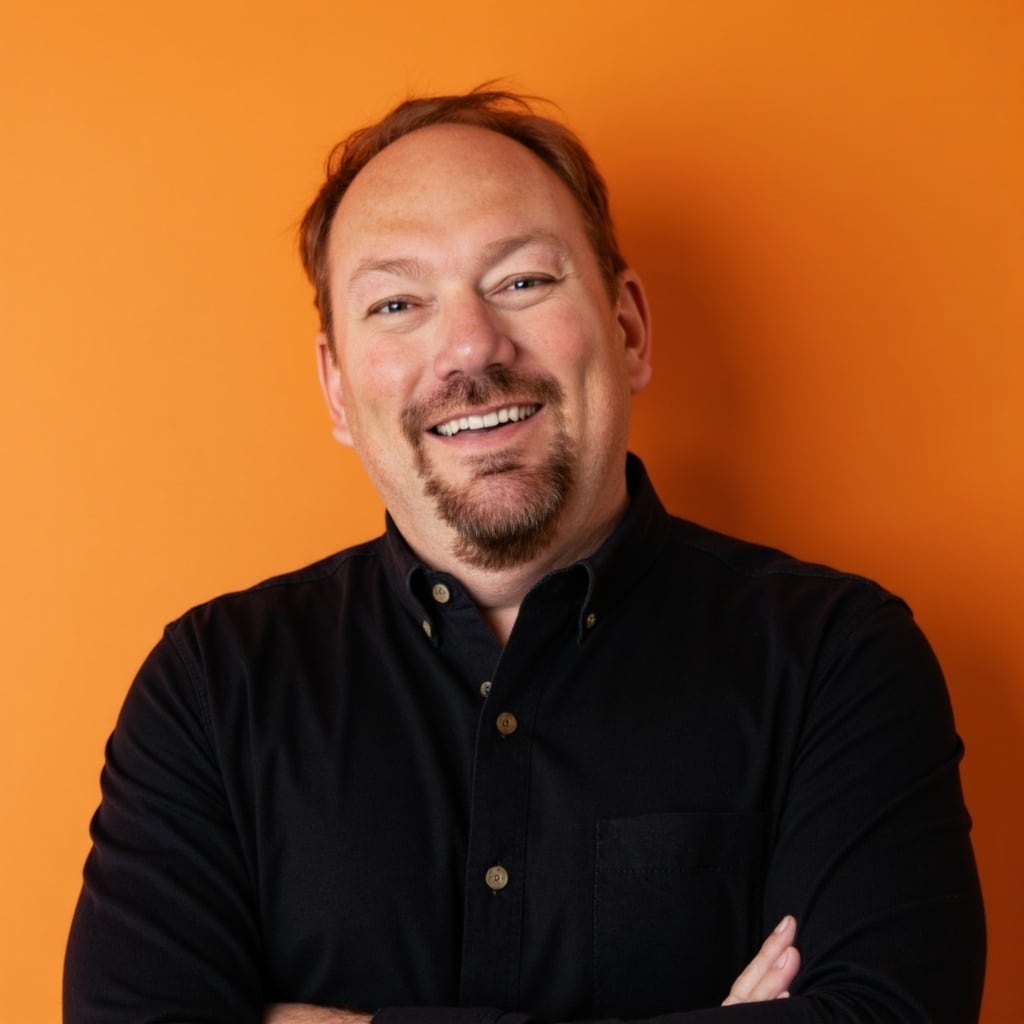 A man in a black button up dress shirt stands in front of an orange background.