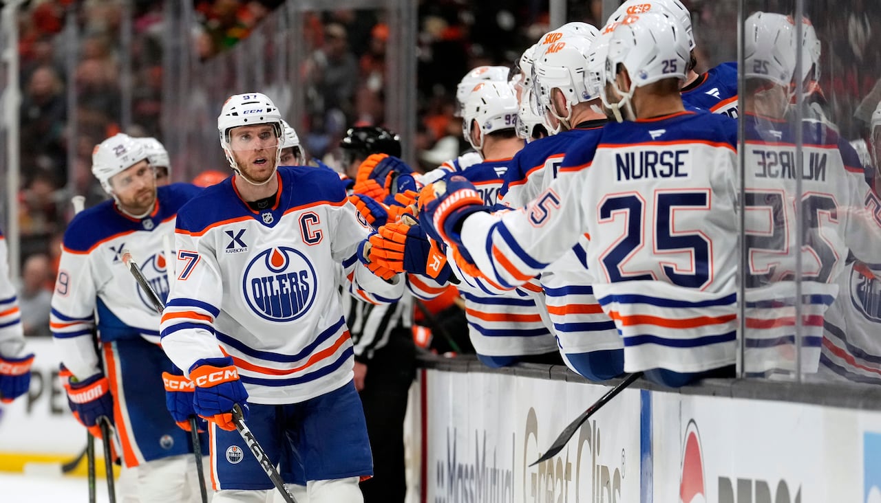 A line of men's hockey players in white-blue-and-orange uniforms fist-bumps their teammates on the bench after scoring a goal.