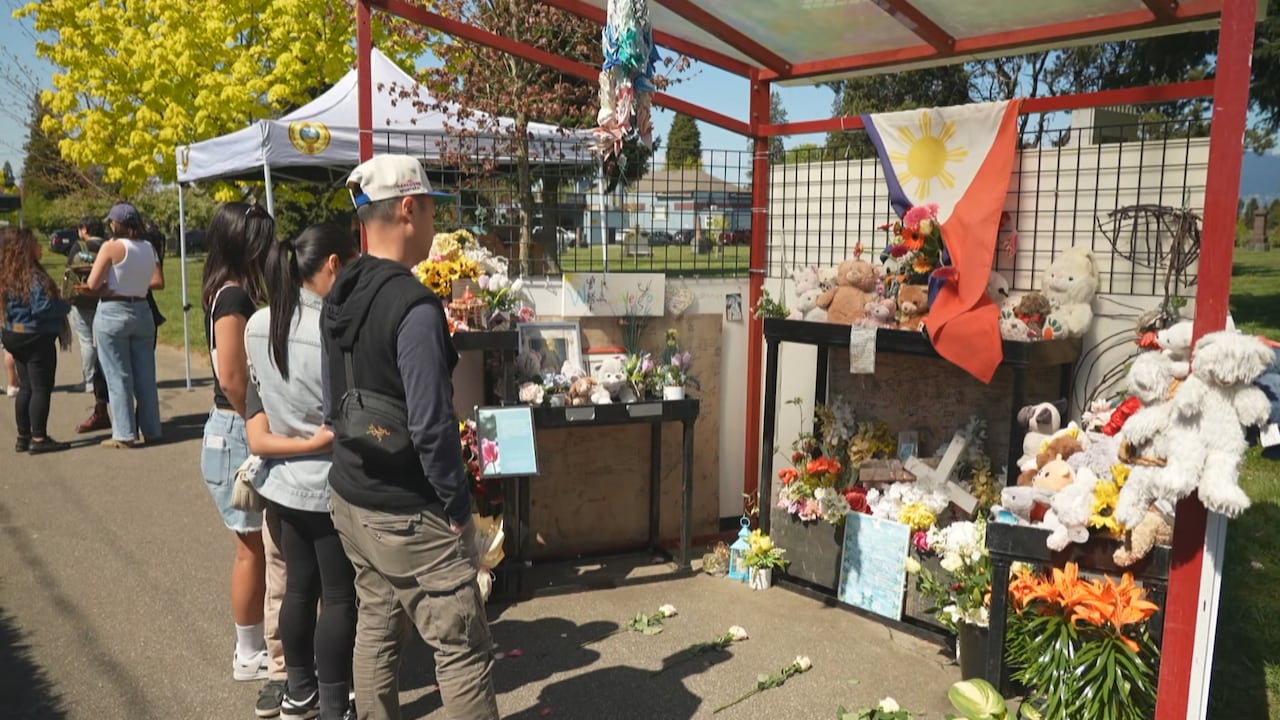 Several people stand facing a memorial display with flowers, stuffed animals and a flag under a small structure.