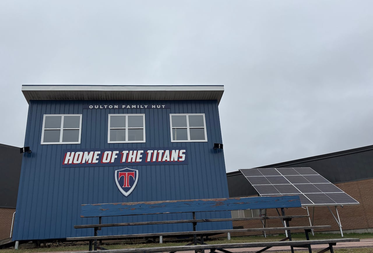 A blue sports building sitting adjacent a football field at a school. On the right, are 16 solar panels and in front of the building are bleachers.