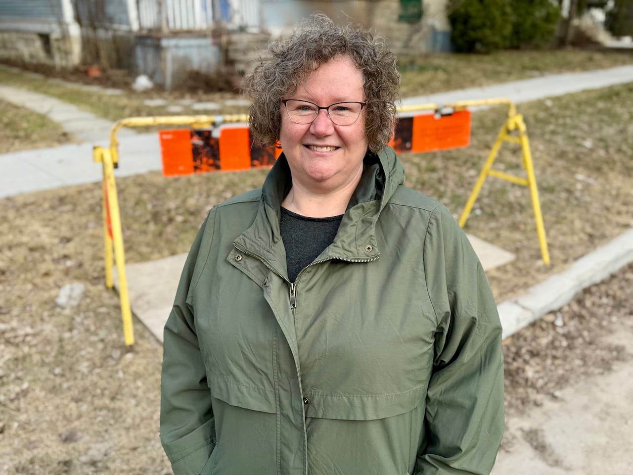 A woman stands outside and smiles. Just behind her on a grassy boulevard is a barricade and sheet of plywood.
