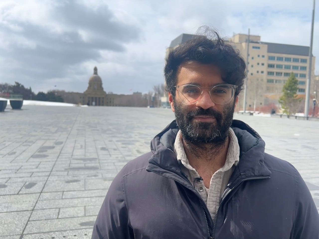 A man with a beard and glasses looks at the camera. He is standing in front of the Alberta legislature.