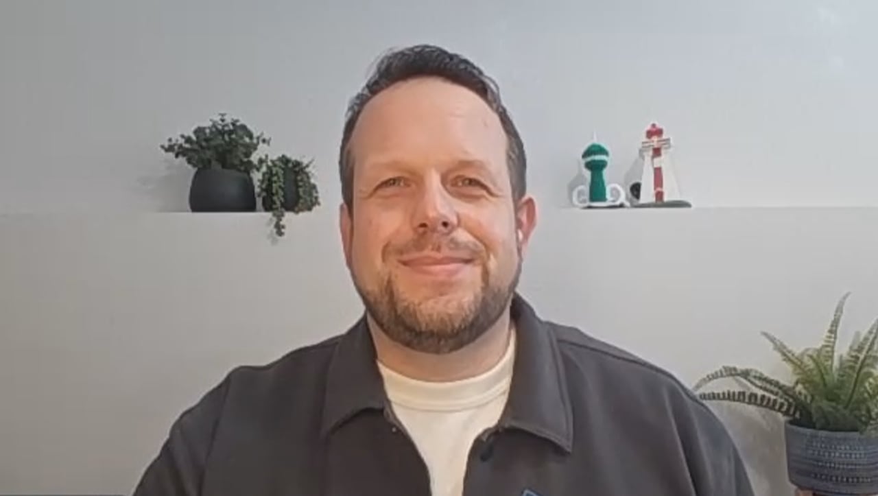 A man wearing a grey sweater and a white t-shirt looks at the camera. He is sitting in front of plants.