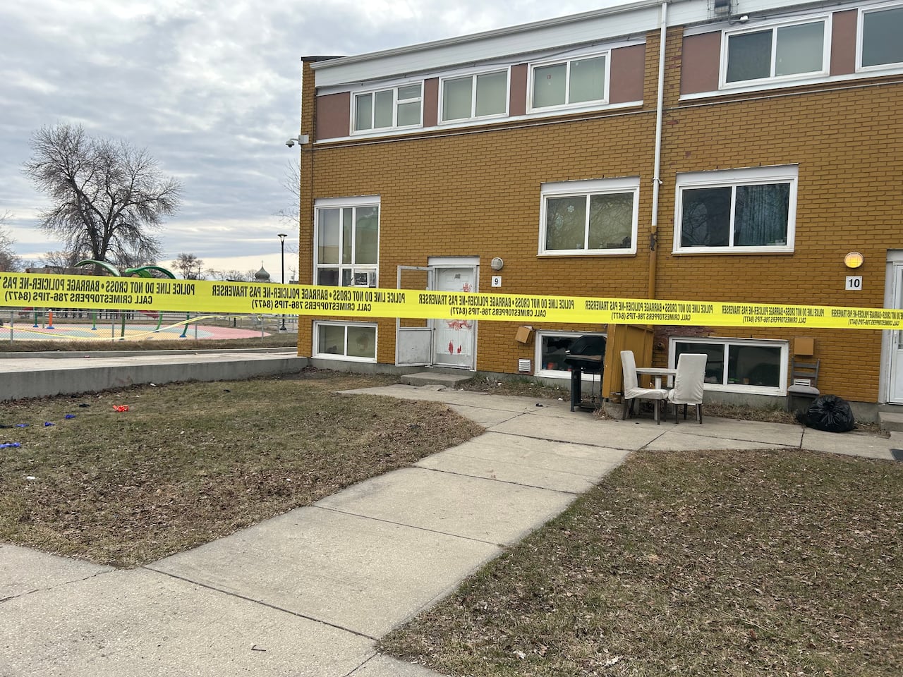 Wide shot of a building. Police tape is in front and blood can be seen on the door.