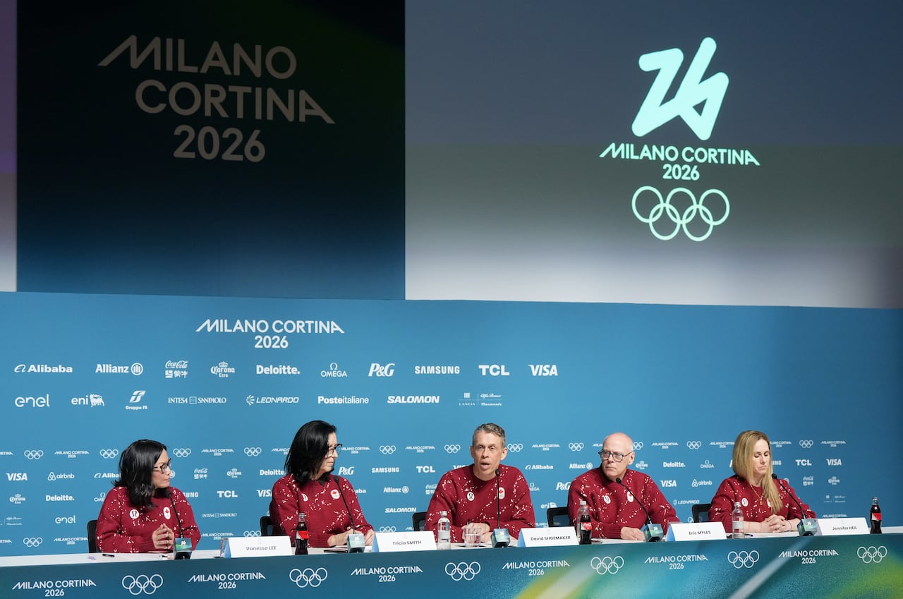 Several people sit at a long table underneath an Olympic logo.