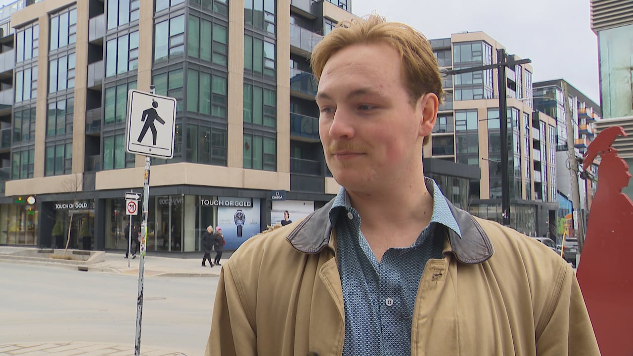 A man wearing a collared shirt and a light jacket has a neutral expression. He is standing on a downtown sidewalk.