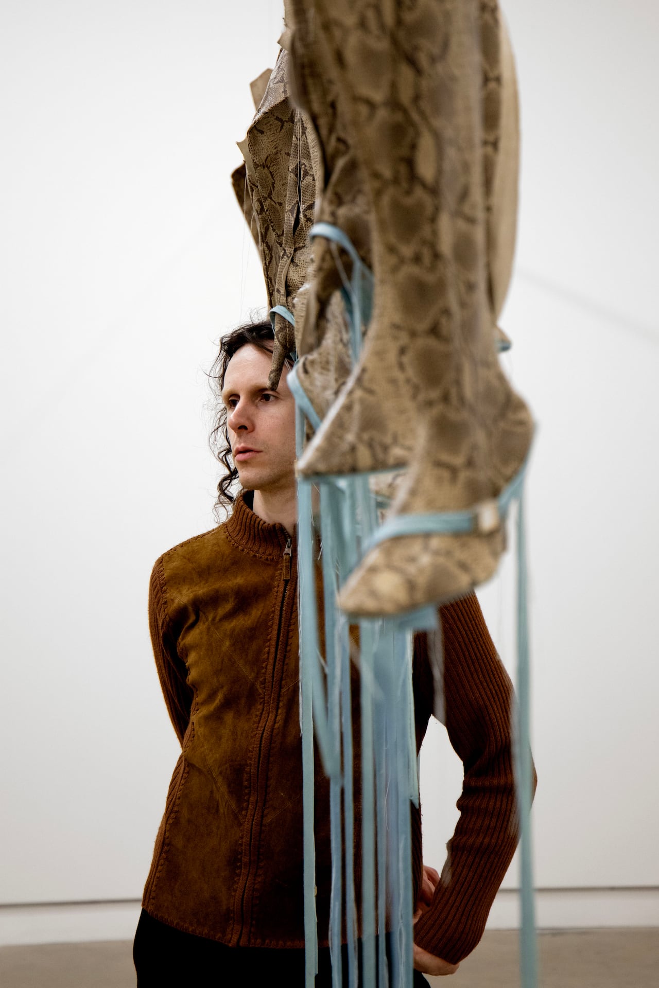 A person with long dark hair and a brown zip-up sweater stands behind a row of snakeskin boots with long blue ribbons hanging from above.