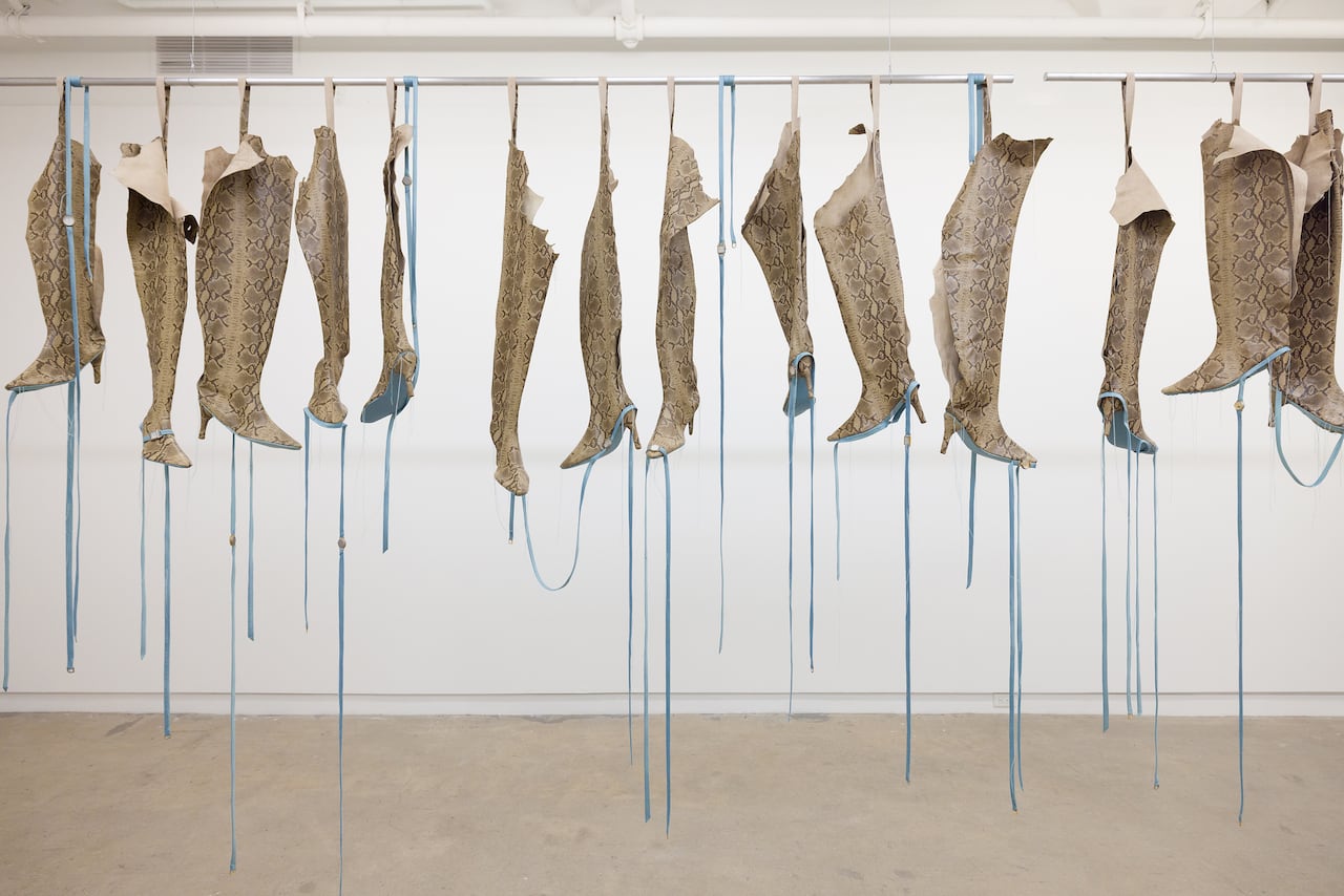 In a room with white walls, a rod spans the space. From it, snakeskin boots with long blue ribbons are hung.