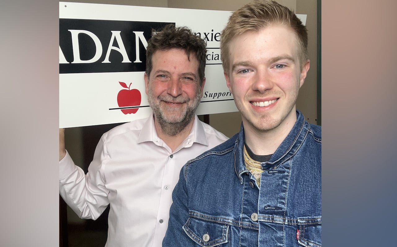 A man with short, dark hair and a salt-and-pepper beard smiles to the camera, standing next to a younger man wearing a dark jean jacket.