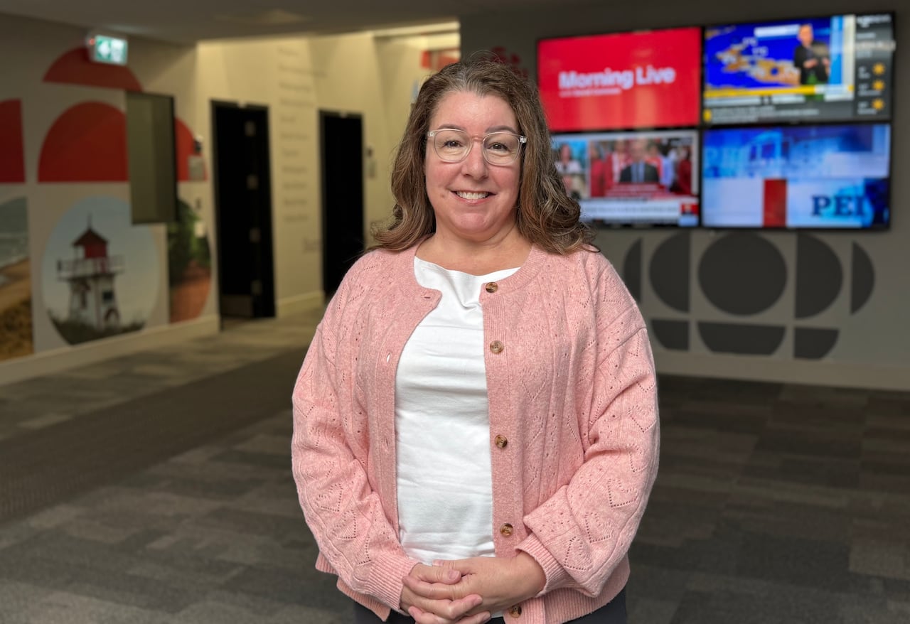 A smiling woman with brown hair and glasses wearing a pink cardigan stands in a modern office lobby featuring media screens and a lighthouse mural.