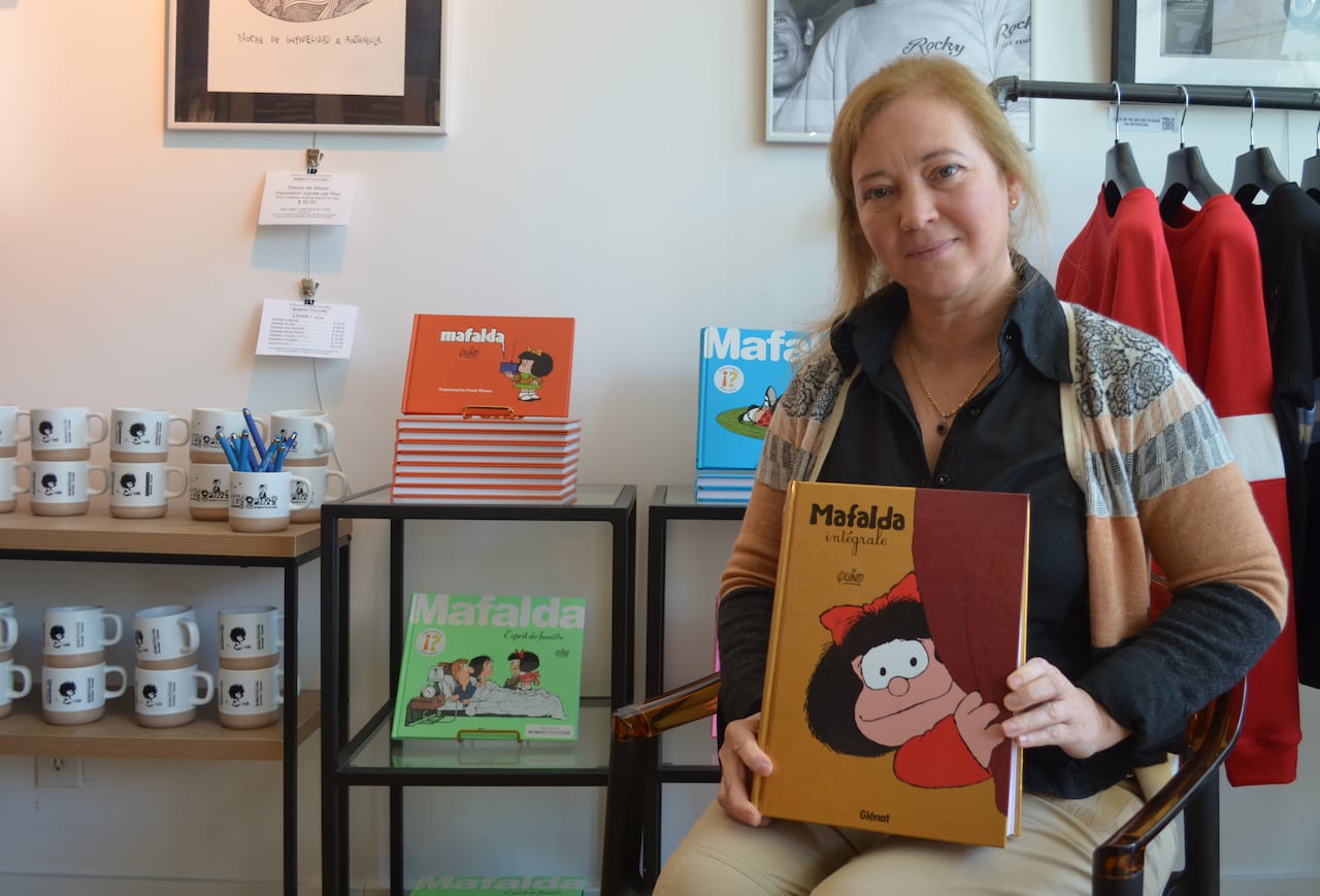 A woman seated in front of Mafalda merch poses with a book of Mafalda