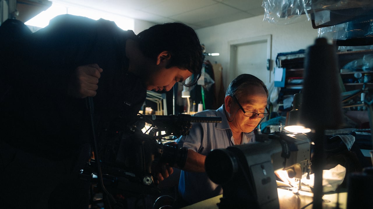 Inside a workshop, a videographer films an tailor who is focused on working at his sewing machine.