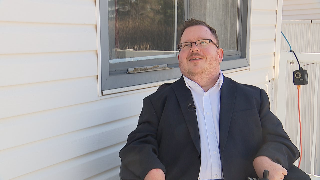 A man in an electric mobility chair sits outside of a home. He has glasses and is wearing a white button-down shit and a black suit jacket.