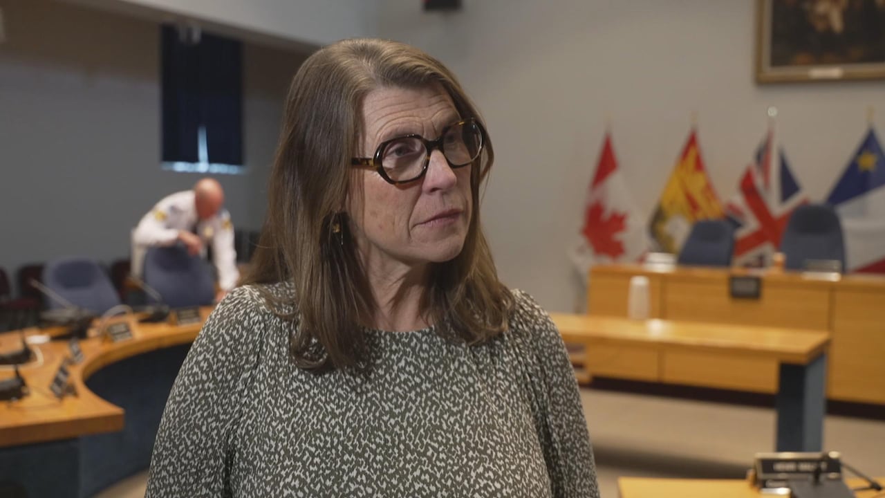 A woman with glasses looks to the side while standing in a council chamber.