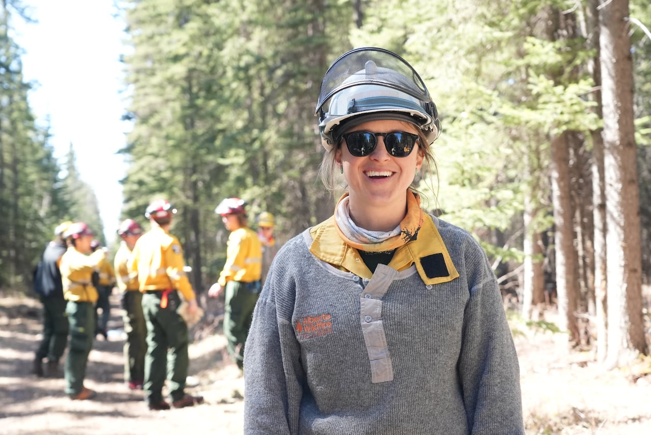 Woman smiles at camera in forest and other firefighter personnel stand in background.