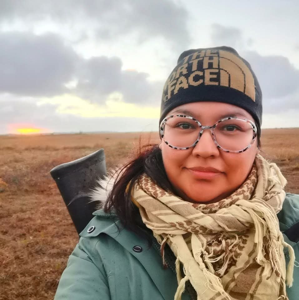 A picture of a woman wearing a tuque and parka with a rifle slung over her shoulder and a flat grassy background receding to a grey sky