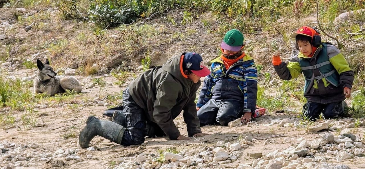 Three boys wearing ball caps and outdoor clothes play on the ground with rocks while a dog looks on