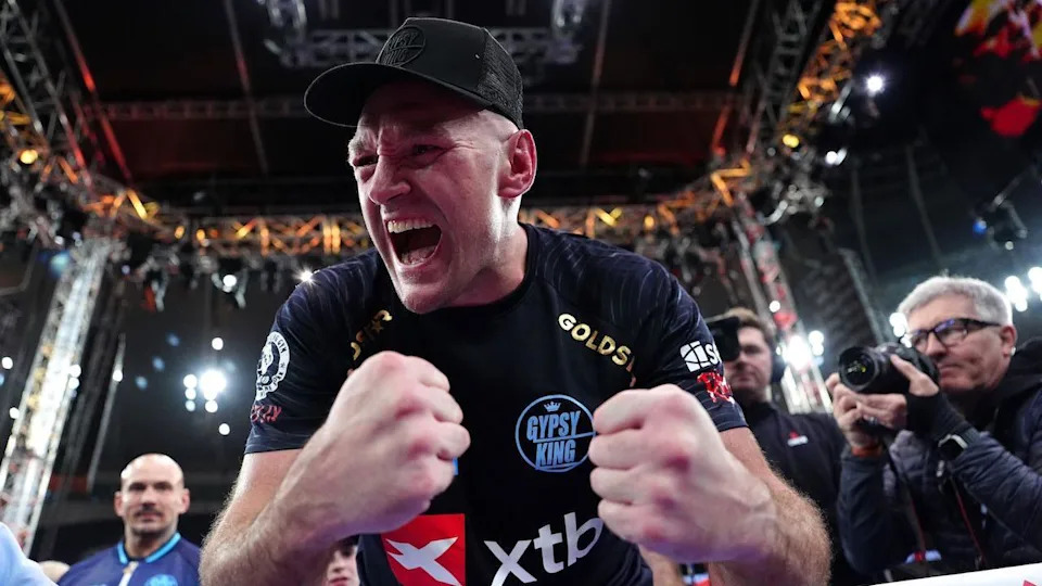 Tyson Fury celebrates, he wears a black t-shirt and cap as he clenches his fists in a sports arena, people take pictures of him in the background.