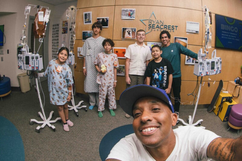 A group of children in hospital attire and adults pose and smile together in a hospital room, with one man taking a selfie. Medical equipment and cheerful decorations are visible around them.
