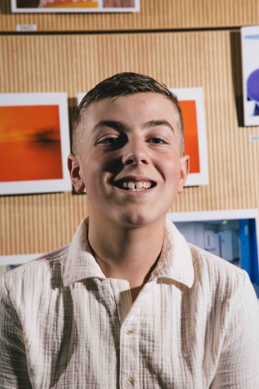 A smiling teenage boy with short hair wearing a textured white shirt stands in front of a wall displaying colorful framed photos or artwork.
