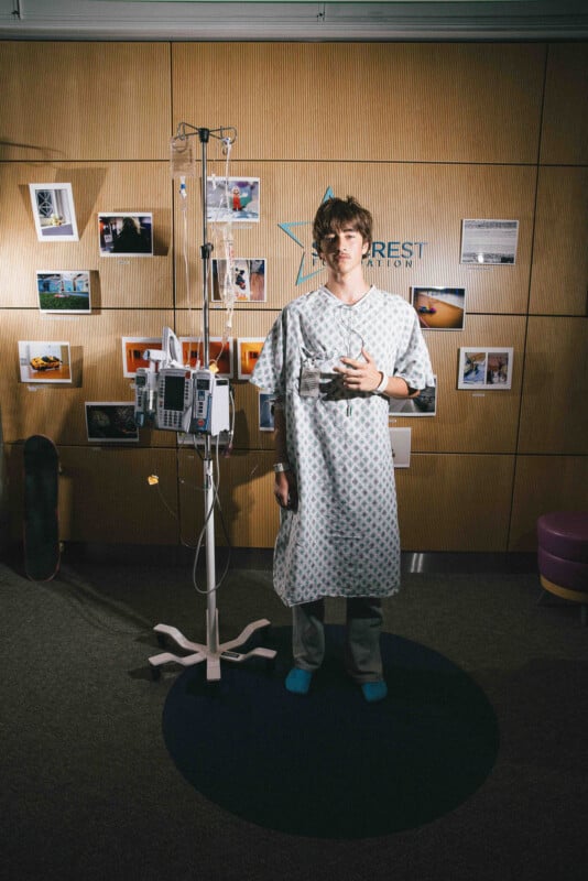 A young man in a hospital gown stands indoors next to an IV pole with medical equipment, holding a cup. He is barefoot on one foot, wearing socks, and stands in front of a wall with photos and a skateboard to his left.
