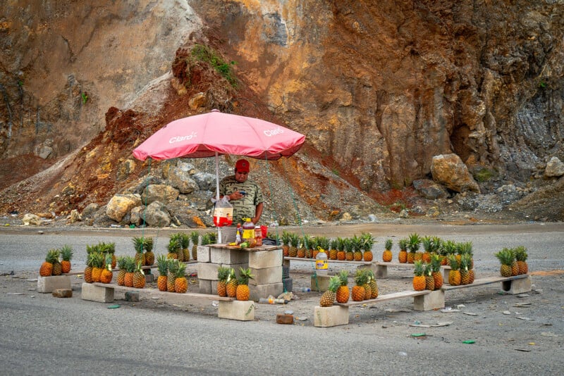 A street vendor stands under a pink umbrella selling pineapples arranged neatly on concrete blocks in a square, with a rocky hillside in the background.