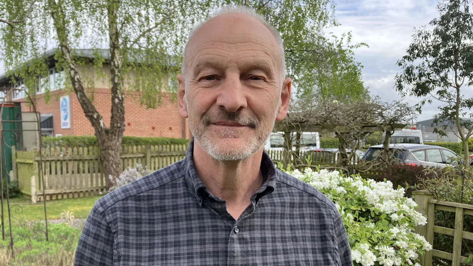 A man is smiling at the camera. He has grey hair and a grey beard. He is wearing a grey checked shirt and standing in a garden.