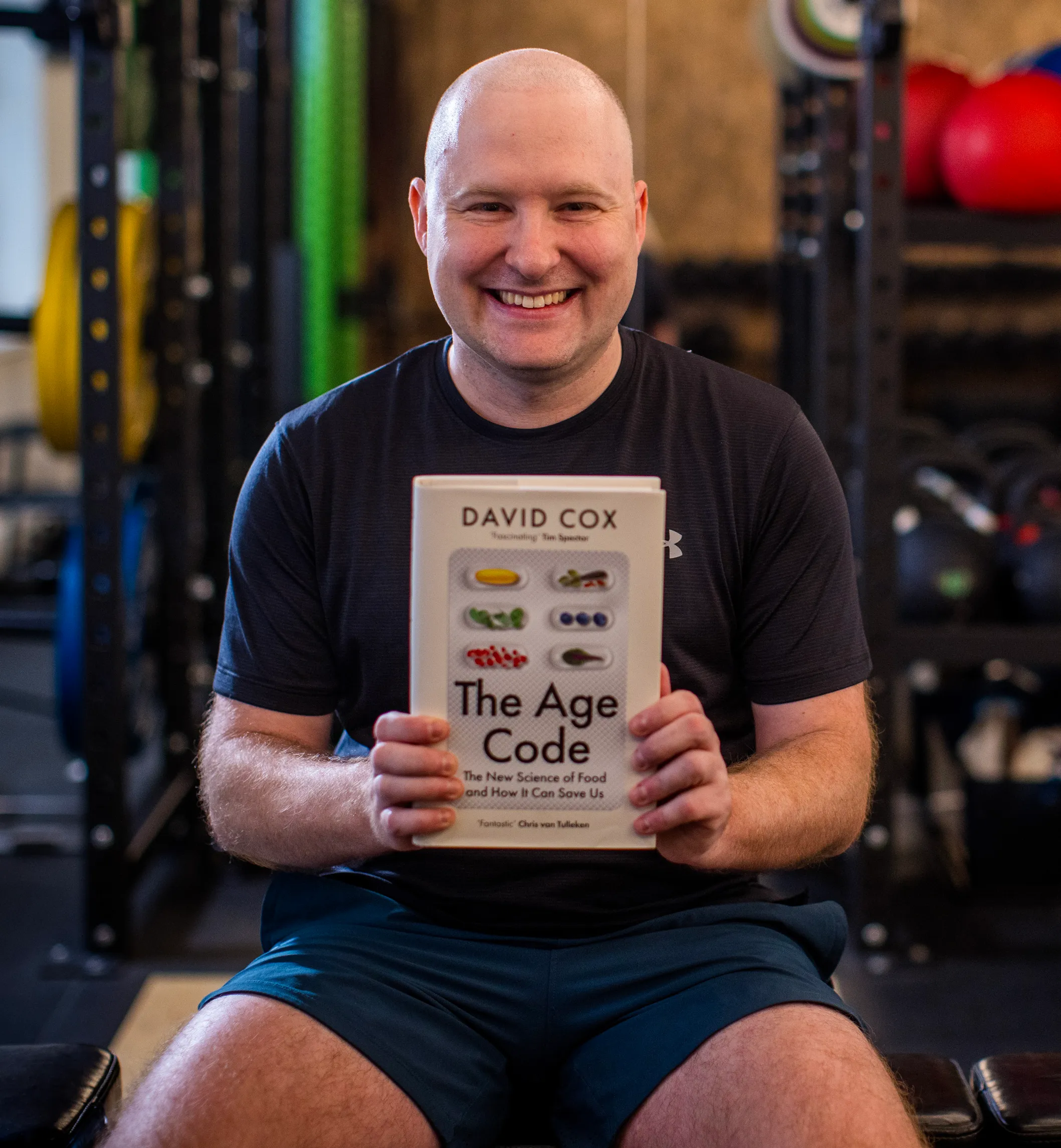 David Cox, a bald man in a dark t-shirt, smiles while holding his book "The Age Code" in a gym.
