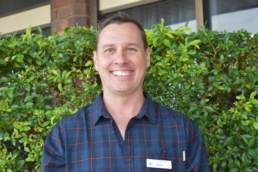 A head-and-shoulders mid shot of a man looking directly at the camera in front of a green hedge. 