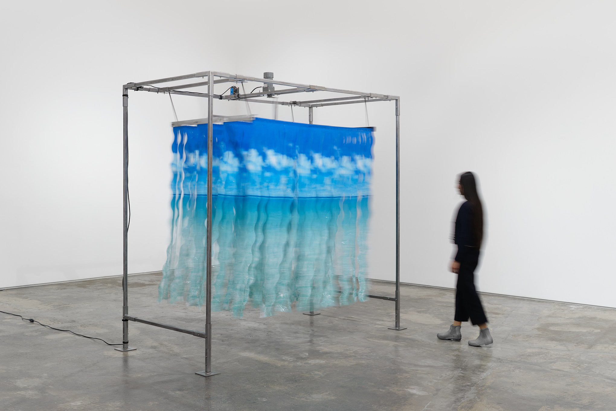 Shredded shower curtains featuring a beach scene hanging from a metal frame in a gallery.