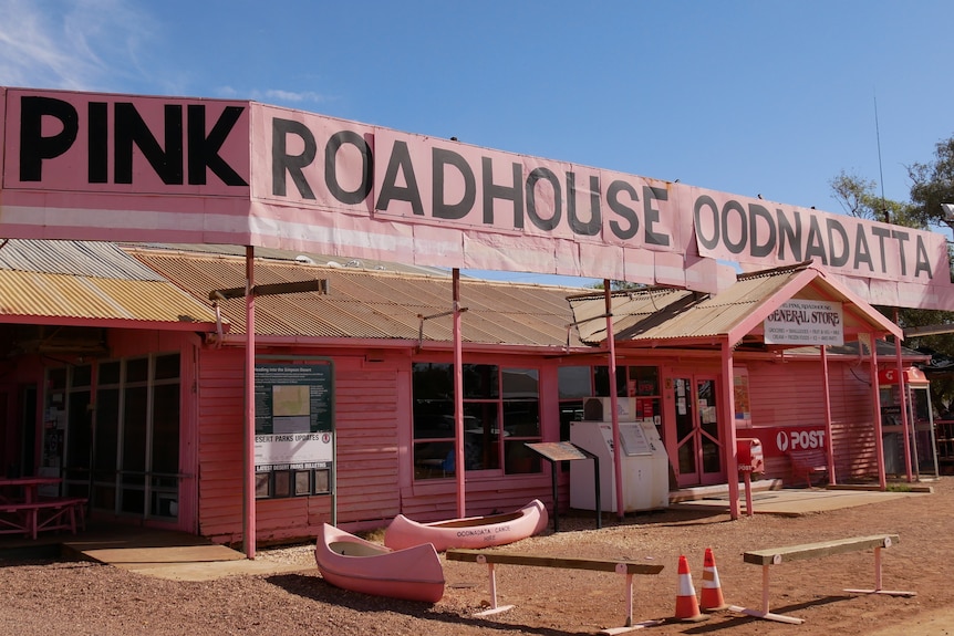 A large building exterior panted pale pink with two pink canoes outside. The sign reads 'PINK ROADHOUSE OODNADATTA' in black.