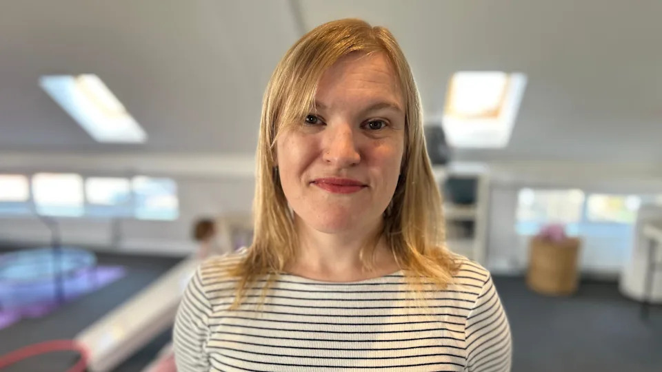Claire looking into camera with children's toys behind her. She has shoulder-length blonde hair and is wearing a black and white striped top.