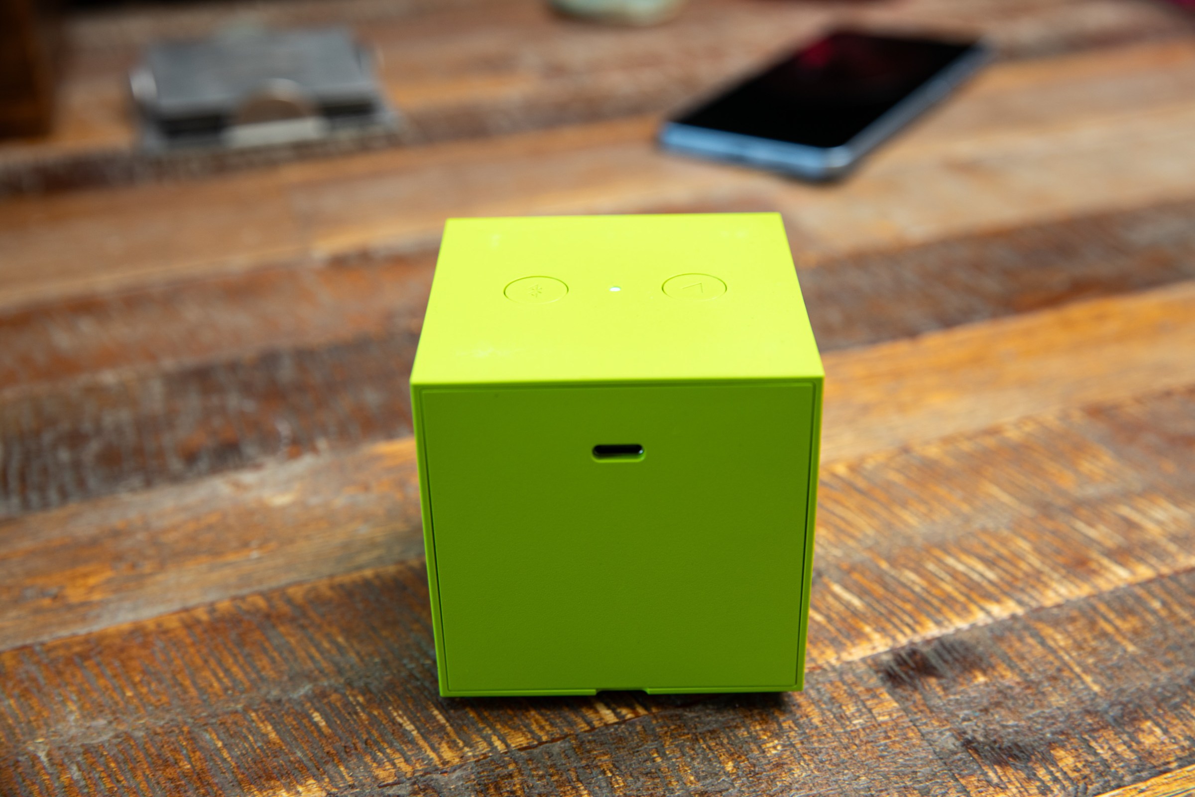 The back of a green Ikea Kallsup speaker on a wooden table, showing its USB-C charge port and two buttons.