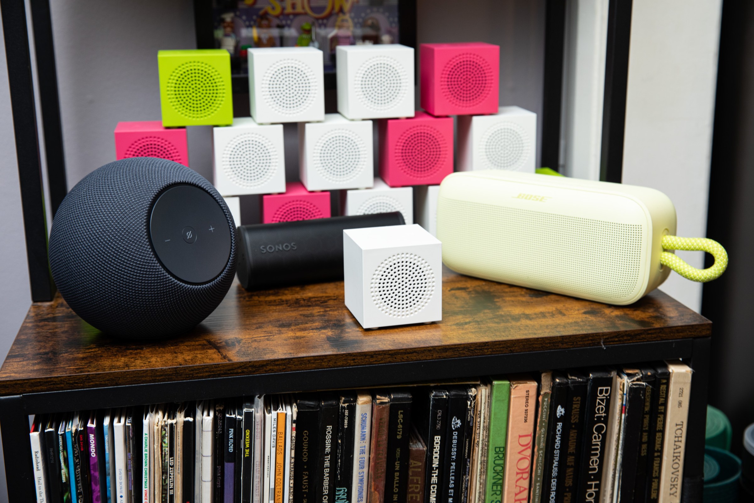 A stack of green, white, and pink Ikea Kallsup speakers behind an Amazon Echo Studio, Sonos Roam 2, Bose SoundLink Plus, and single white Kallsup speaker on a wooden vinyl record shelf.